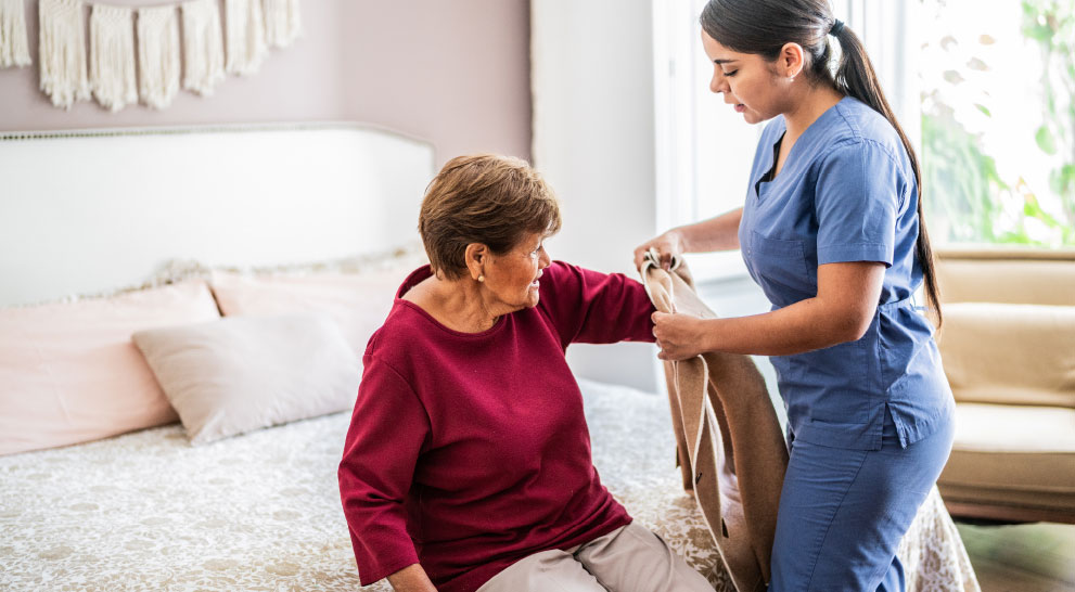 assisted_living_rehab_image (2) A nurse assists a senior woman put on a sweater.